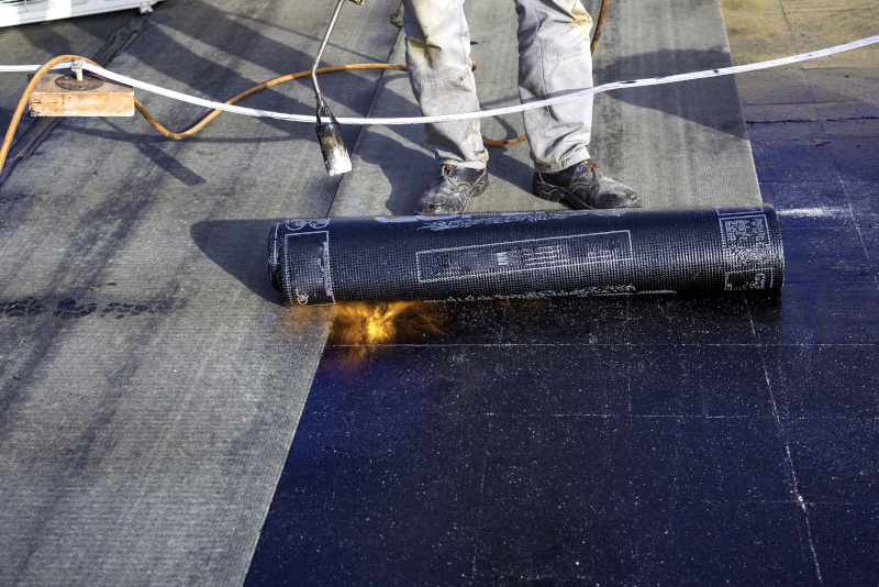 Worker preparing part of bitumen roofing felt roll for melting by gas heater torch flame; commercial roofing