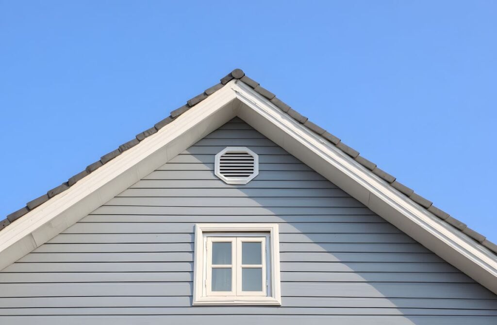 House exterior detail featuring peak of gable roof with gray vinyl siding white fascia and soffit trim rectangular window and round attic vent